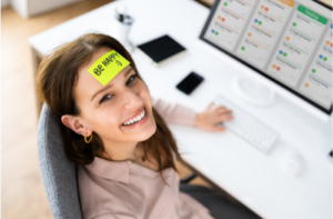 Une femme travaille à sur son ordinateur. Elle sourit, et porte un post-it sur le front avec l'inscription "be happy"